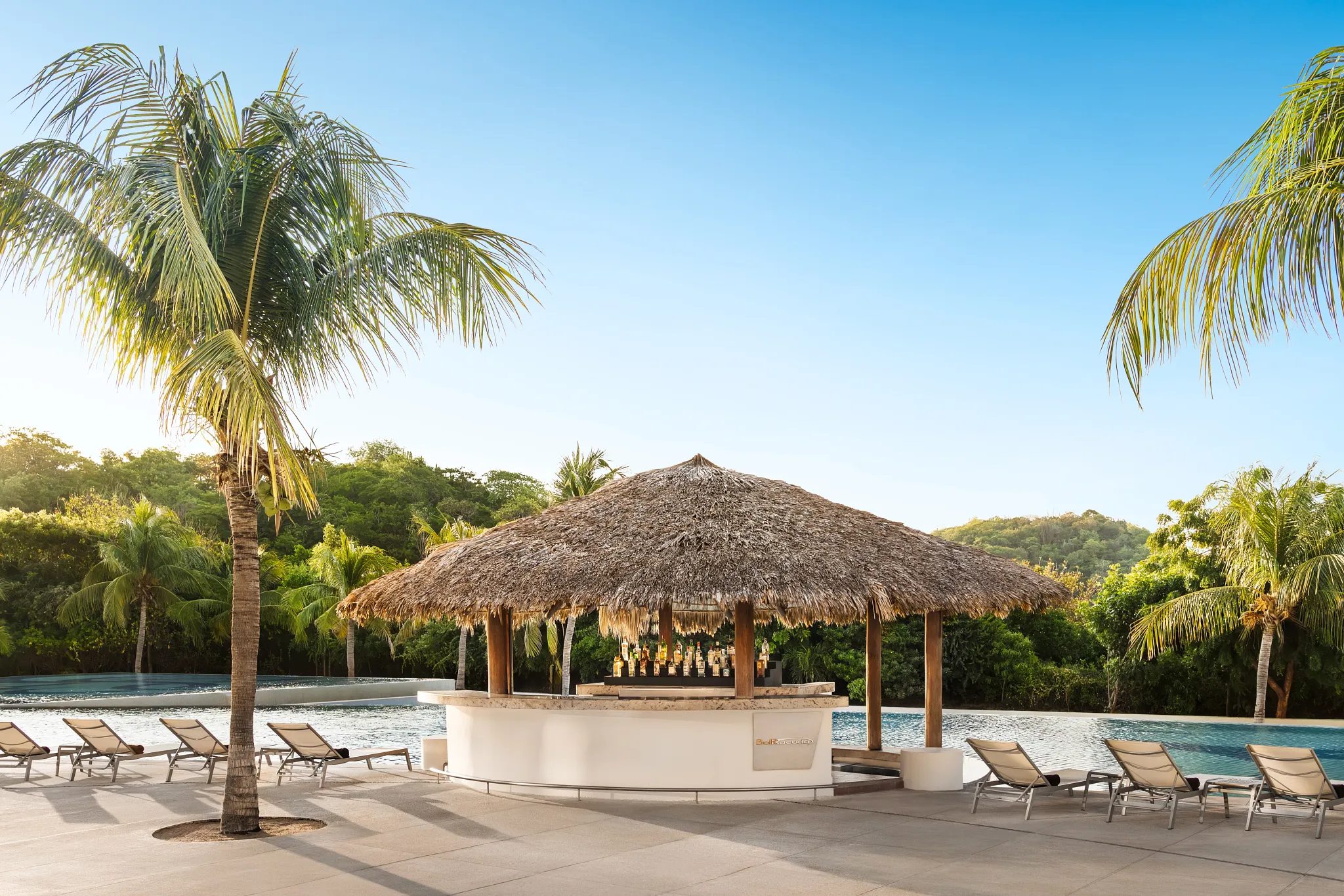 Barracuda pool and beach bar at Secrets Huatulco Resort & Spa featuring a thatched-roof bar, tropical cocktails, and palm-lined poolside setting.