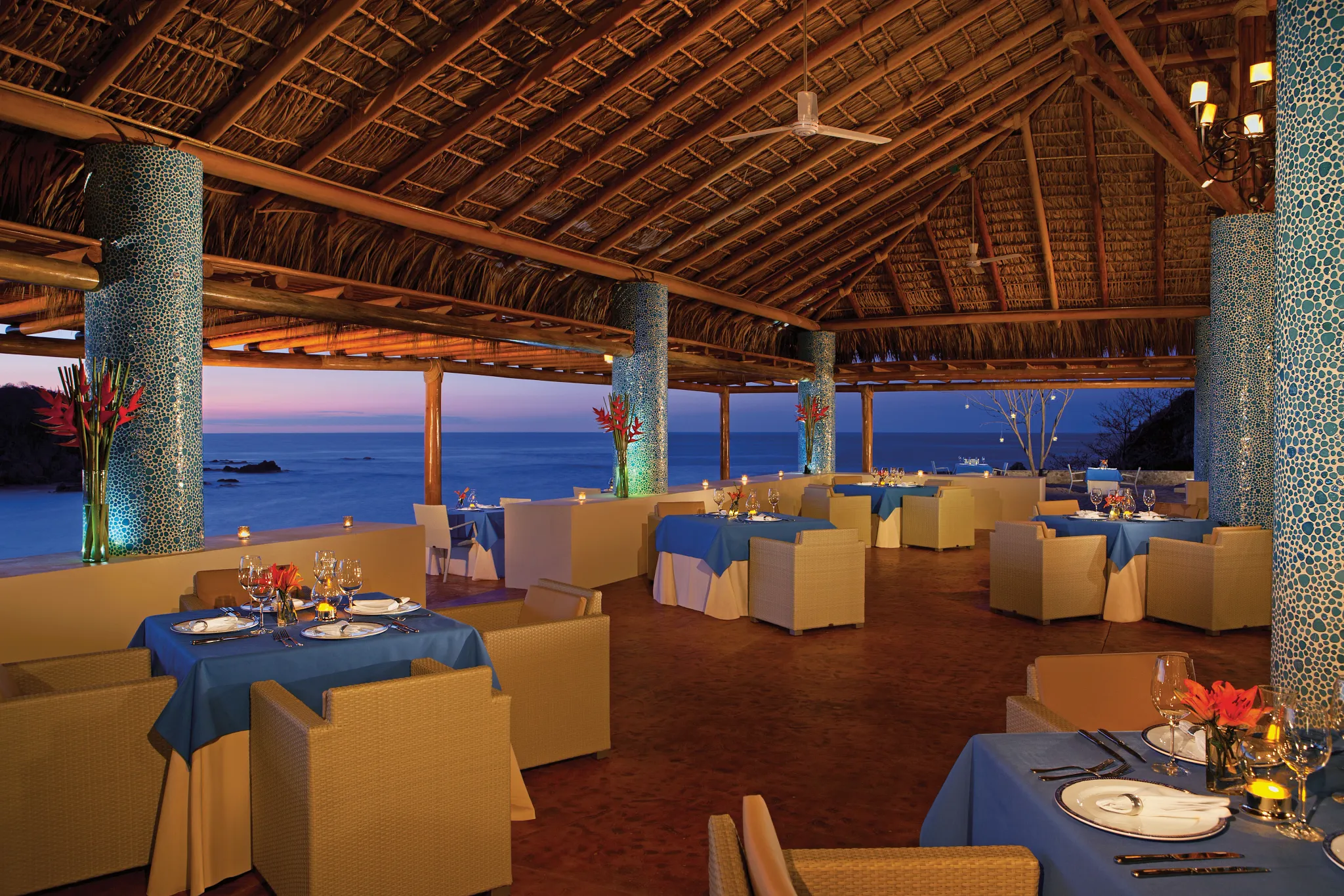 Castaways restaurant at Secrets Huatulco Resort & Spa featuring open-air oceanfront dining, thatched roof design, and elegant table settings at sunset.