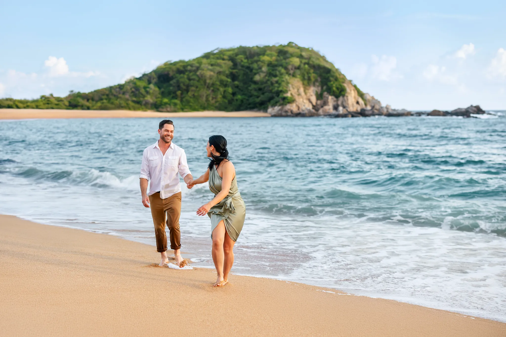 Romantic couple walking hand in hand along the sandy beach at Secrets Huatulco Resort & Spa with lush hills in the background.