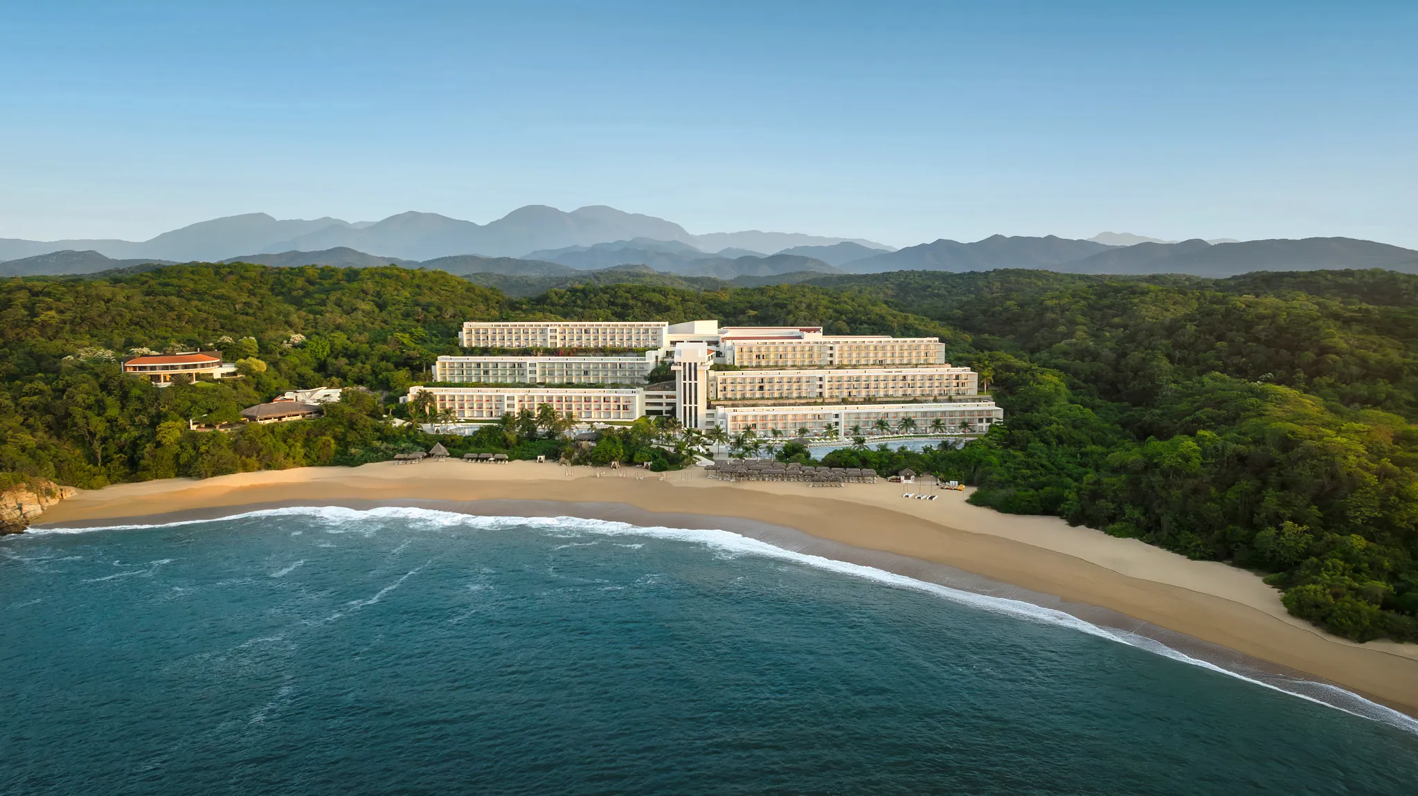 Aerial view of Secrets Huatulco Resort & Spa beachfront with jungle hills and mountain views behind.