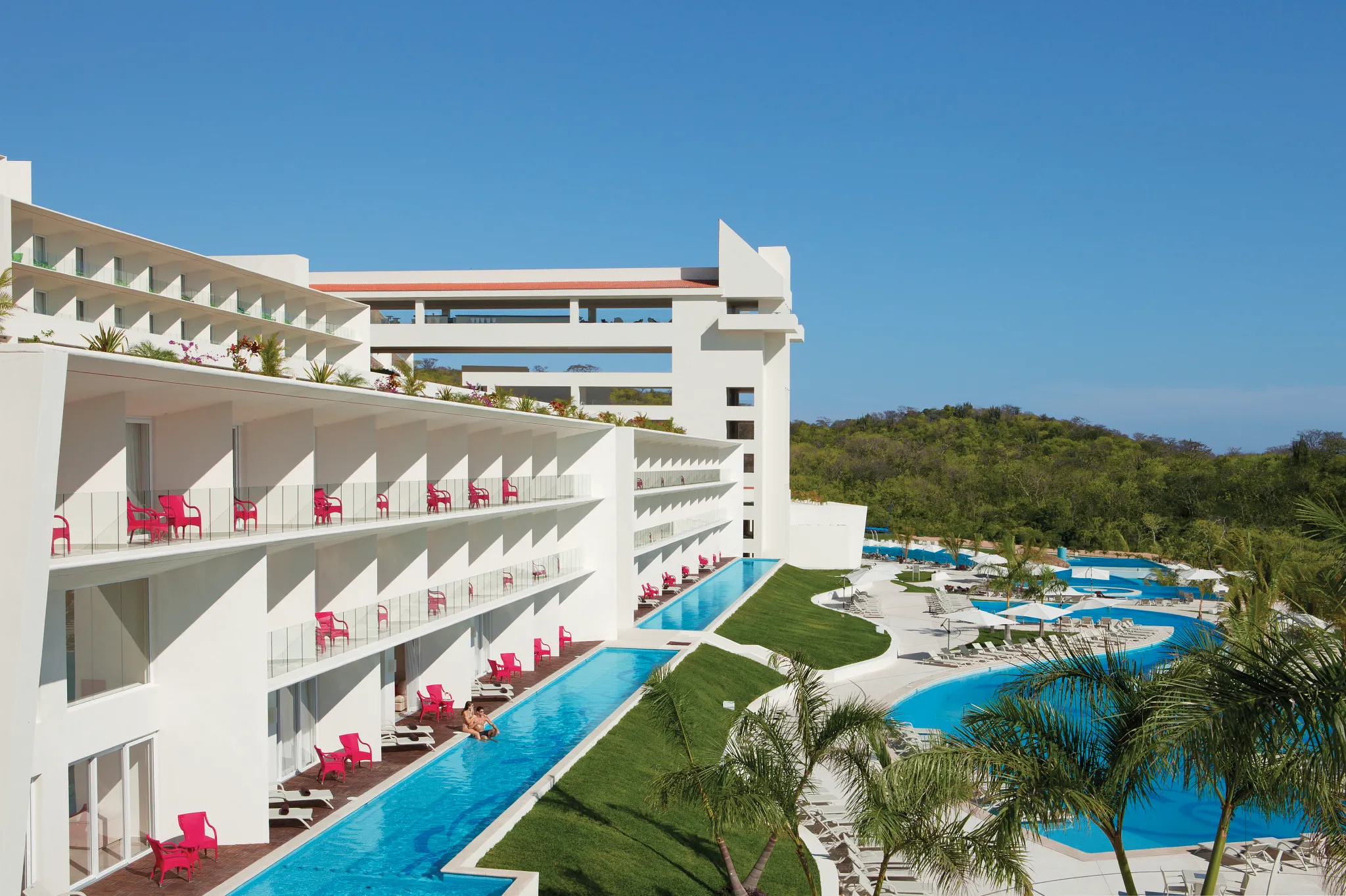 Modern resort buildings and infinity-style pools at Secrets Huatulco Resort & Spa surrounded by lush hills and ocean views.