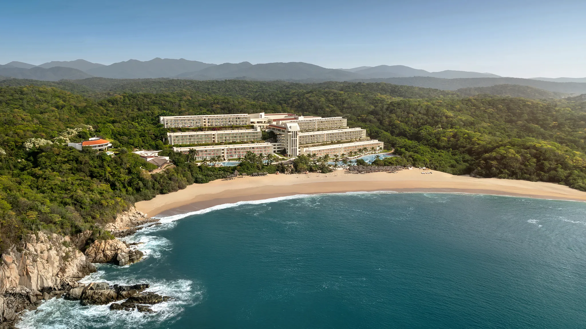 Aerial view of Secrets Huatulco Resort & Spa along a quiet bay surrounded by jungle and mountains.